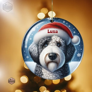 May include: A round ceramic ornament featuring a fluffy black and white dog wearing a red and white Santa hat. The name Luna is printed in red text above the dog. The background is a dark blue winter scene with white snowflakes and a gold string.