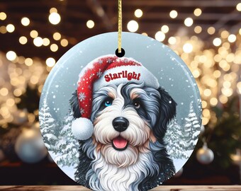 May include: Round Christmas ornament featuring a dog wearing a Santa hat with the word "Starlight". The dog has black, white, and brown fur, with one blue eye. The background shows a snowy scene with Christmas trees.