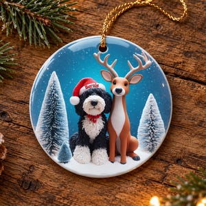 May include: A round ceramic Christmas ornament featuring a black and white dog wearing a red Santa hat with the name Bernie printed on it, sitting next to a brown deer in a snowy forest with pine trees under a blue sky.