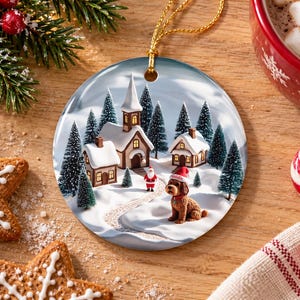 May include: A round ceramic Christmas ornament featuring a snowy village scene with a church, houses, pine trees, a small Santa Claus figure, and a brown dog wearing a red Santa hat with the name Goldie printed on the brim.
