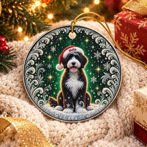 May include: A round ceramic Christmas ornament featuring a black and white dog wearing a red Santa hat with the name Stella printed on the brim. The dog is centered against a dark green background with white snowflakes and ornate silver scrollwork.