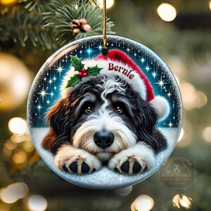 May include: A round Christmas ornament featuring a Bernedoodle dog wearing a Santa hat with the name "Bernie". The ornament has a snowy background with sparkling stars and holly berries. A gold string is attached for hanging.