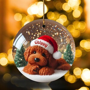 May include: A spherical Christmas decoration showing a brown dog wearing a Santa hat with the name "Chippy". The ornament has a snowy base with small green trees, set against a blurred background of golden lights.