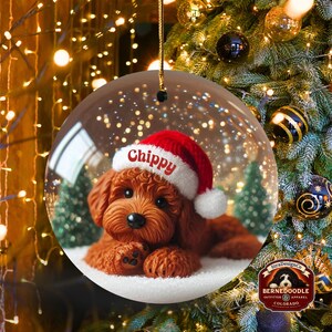 May include: A clear, round Christmas bauble with a brown dog wearing a Santa hat, the name "Chippy" is on the hat. The bauble contains a snowy scene with small green trees. The background is a Christmas tree with lights and decorations.
