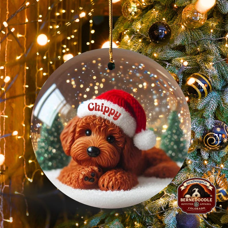 May include: A clear, round Christmas bauble with a brown dog wearing a Santa hat, the name "Chippy" is on the hat. The bauble contains a snowy scene with small green trees. The background is a Christmas tree with lights and decorations.