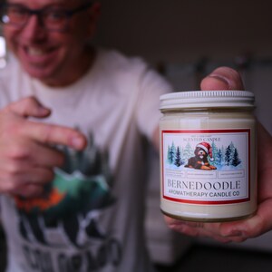Puede incluir: Una vela en tarro de cristal con tapa blanca y etiqueta con un perro Bernedoodle con gorro de Pap&aacute; Noel. La etiqueta tambi&eacute;n incluye el texto "BERNEDOODLE AROMATHERAPY CANDLE CO."
