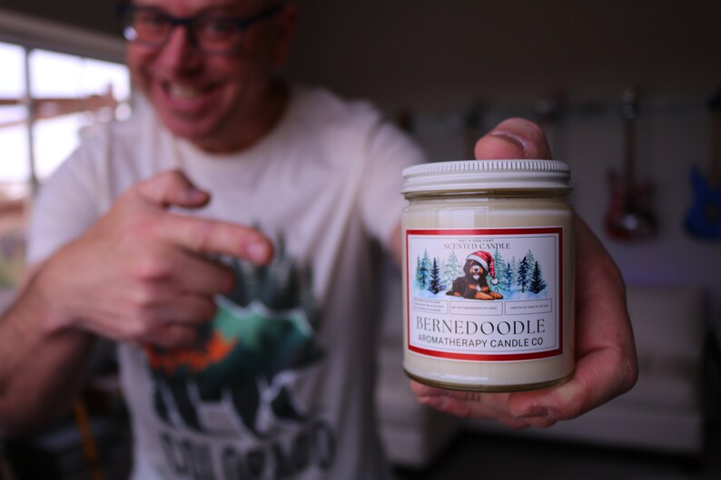 Puede incluir: Una vela en tarro de cristal con tapa blanca y etiqueta con un perro Bernedoodle con gorro de Pap&aacute; Noel. La etiqueta tambi&eacute;n incluye el texto "BERNEDOODLE AROMATHERAPY CANDLE CO."