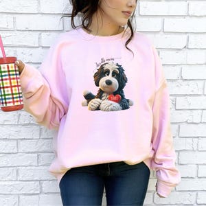 A woman wearing a Bernedoodle sweatshirt with cute design on the front and the back.  The front features a bernedoodle dog in claymation design holding a heart with his name, across the top of his head the words, "Doodle Mom".