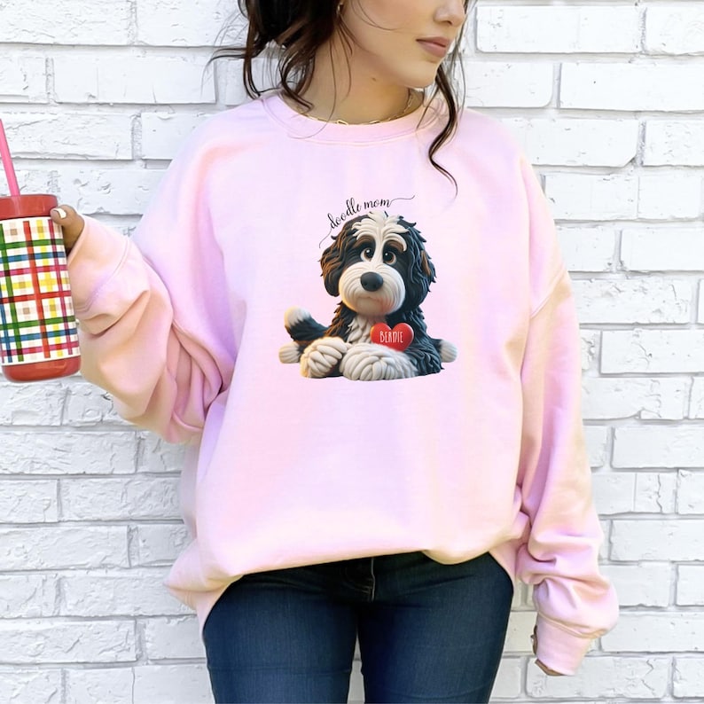 A woman wearing a Bernedoodle sweatshirt with cute design on the front and the back.  The front features a bernedoodle dog in claymation design holding a heart with his name, across the top of his head the words, "Doodle Mom".