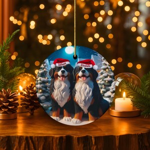 Op de afbeelding: Een ronde kerstversiering met twee Berner Sennenhonden met kerstmutsen. De honden worden afgebeeld in een winterse sc&egrave;ne met besneeuwde bomen. De namen "Edison" en "Einstein" staan op de mutsen. De versiering hangt aan een gouden draad.