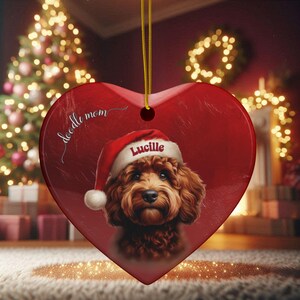 May include: Heart-shaped Christmas ornament with a red background and a portrait of a brown dog wearing a Santa hat. The ornament has the text "doodle mom" and the name "Lucille". A gold string is attached for hanging.