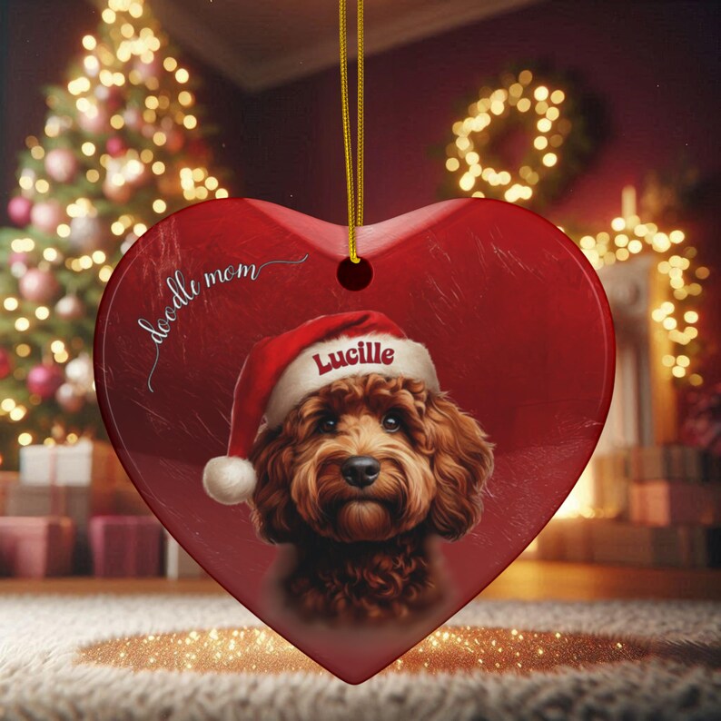 May include: Heart-shaped Christmas ornament with a red background and a portrait of a brown dog wearing a Santa hat. The ornament has the text "doodle mom" and the name "Lucille". A gold string is attached for hanging.