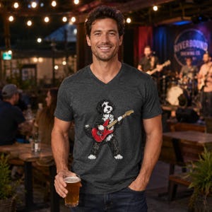 May include: A charcoal gray V-neck t-shirt featuring a graphic illustration of a black and white dog playing a red electric guitar. The shirt has short sleeves and a heathered texture, showcasing a detailed print of the canine musician.