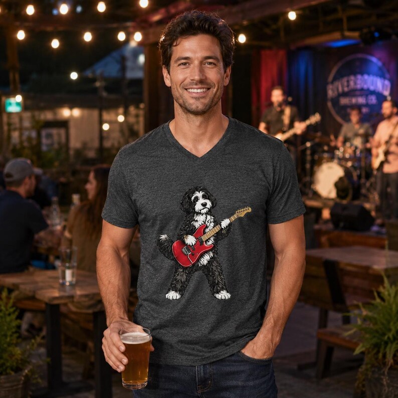 May include: A charcoal gray V-neck t-shirt featuring a graphic illustration of a black and white dog playing a red electric guitar. The shirt has short sleeves and a heathered texture, showcasing a detailed print of the canine musician.