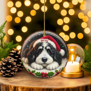 May include: Round Christmas ornament featuring a black and white dog wearing a Santa hat with the name "Bernie". The ornament has a gold string for hanging and is decorated with holly and berries. Festive holiday decor.