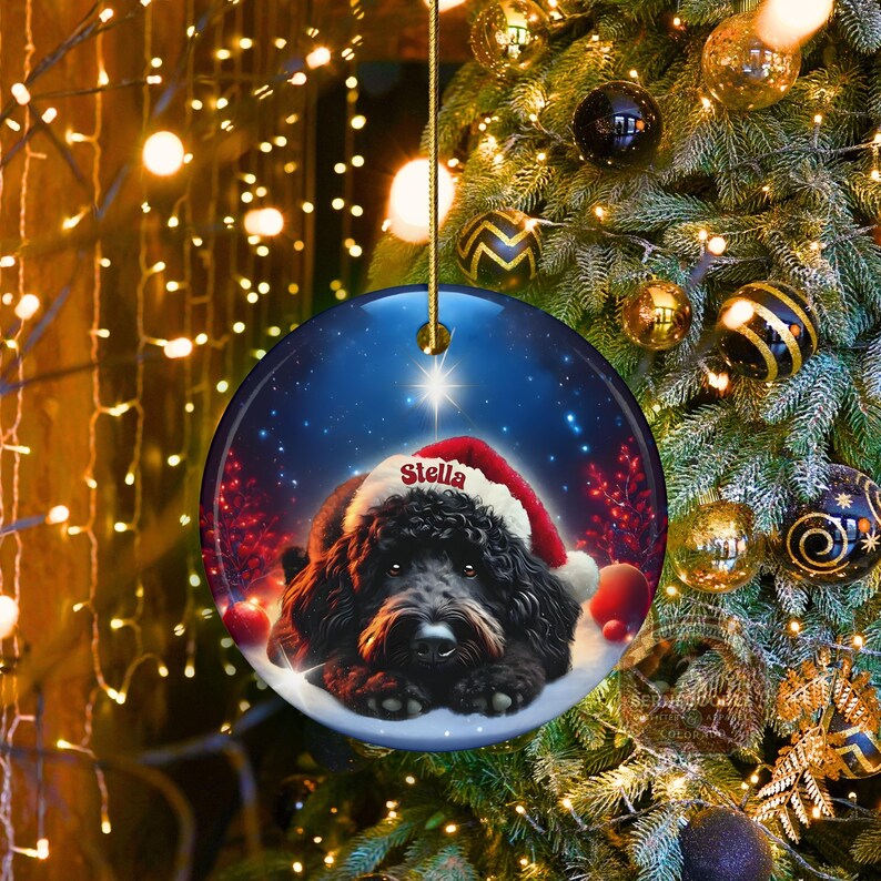 May include: Round Christmas ornament featuring a black dog wearing a Santa hat, with the name "Stella" on the hat. The ornament has a blue background with stars and is hanging from a gold string. Festive holiday decor.