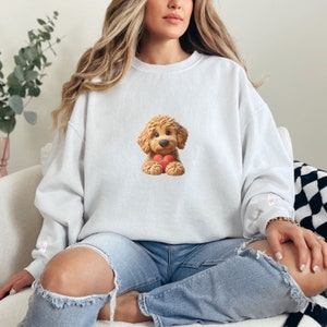 May include: White sweatshirt featuring a cartoon illustration of a brown dog holding a red heart. The dog has a fluffy appearance and is centered on the front of the sweatshirt. The sleeves have a small pink design. The model is wearing blue jeans.