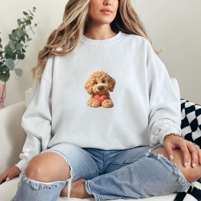 May include: White sweatshirt featuring a cartoon illustration of a brown dog holding a red heart. The dog has a fluffy appearance and is centered on the front of the sweatshirt. The sleeves have a small pink design. The model is wearing blue jeans.
