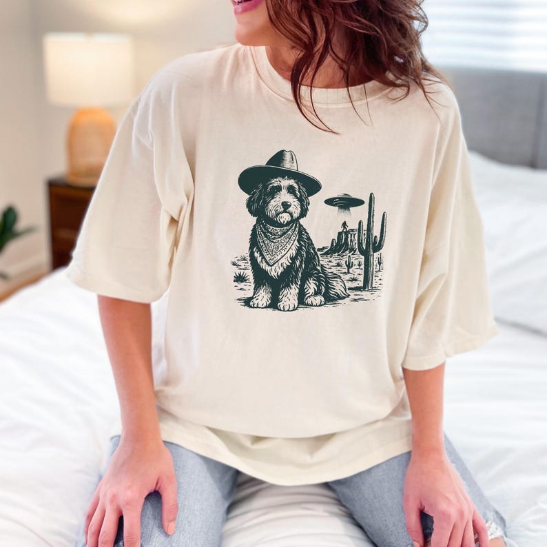 May include: Cream-colored t-shirt featuring a black and white graphic of a dog wearing a cowboy hat and bandana, set against a desert landscape with a UFO. The dog is the central focus of the design.