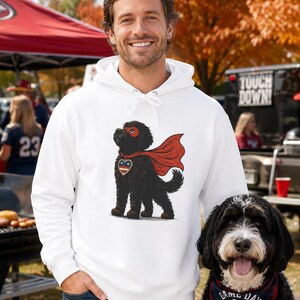 Pu&ograve; includere: Una felpa con cappuccio bianca con la grafica di un cane nero che indossa una maschera e un mantello da supereroe rossi, con un emblema a forma di cuore sul petto. Il cane ha un pelo folto ed &egrave; raffigurato in posizione eretta.