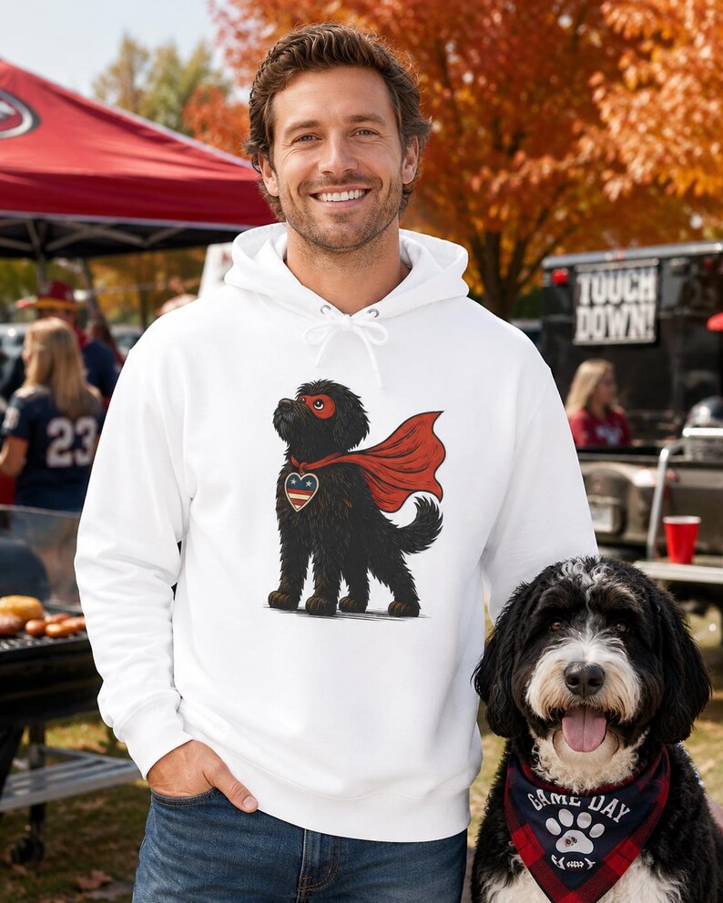 Pu&ograve; includere: Una felpa con cappuccio bianca con la grafica di un cane nero che indossa una maschera e un mantello da supereroe rossi, con un emblema a forma di cuore sul petto. Il cane ha un pelo folto ed &egrave; raffigurato in posizione eretta.