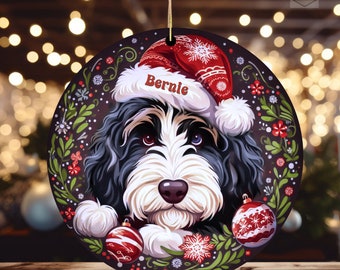 May include: Round Christmas ornament featuring a black, white, and brown dog wearing a red Santa hat with the name "Bernie". The ornament is decorated with red and white snowflakes, red ornaments, and green foliage. A gold string is attached for hanging.