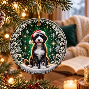 May include: A round ceramic Christmas ornament featuring a black and white Bernedoodle dog wearing a red Santa hat with the name Stella printed on the brim. The dog sits against a dark green background with white snowflakes and gold stars.