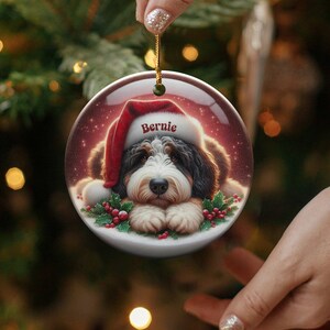Personalize your holiday decor with this charming Bernedoodle Christmas ornament! Featuring a cute Bernedoodle in a festive Santa hat and customizable name, this round ornament adds warmth and joy to any tree. Perfect for dog lovers and unique gifts!