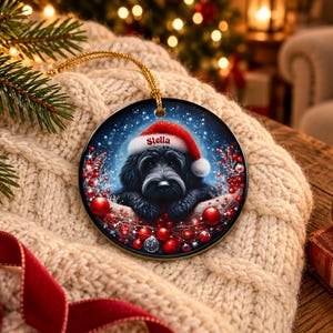 May include: A round ceramic Christmas ornament featuring a black dog wearing a red Santa hat with the name Stella printed on the brim. The dog is surrounded by red berries and ornaments against a dark blue, snowy background with a gold string.