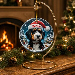 K&ouml;nnte beinhalten: Ein runder Keramikschmuck an einem goldfarbenen Metallst&auml;nder, der einen schwarz-wei&szlig;-braunen Hund mit einer roten Weihnachtsm&uuml;tze zeigt, auf der der Name Bernie in roter Schrift steht, vor einer verschneiten Winterdorfkulisse.