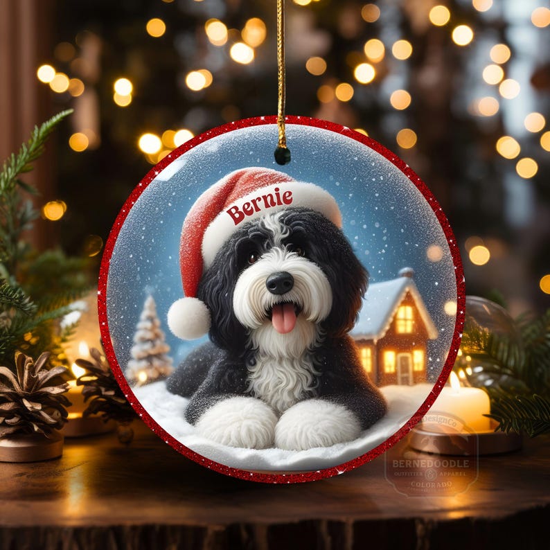 May include: Round Christmas ornament featuring a black and white dog wearing a Santa hat with the name "Bernie". The ornament has a red border and a winter scene with a house and snow. A gold string is attached for hanging.