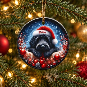 May include: A round ceramic Christmas ornament featuring a black dog wearing a red Santa hat with the name Stella printed on the brim. The dog is surrounded by red berries, red ornaments, and a snowy background, hanging from a gold cord.