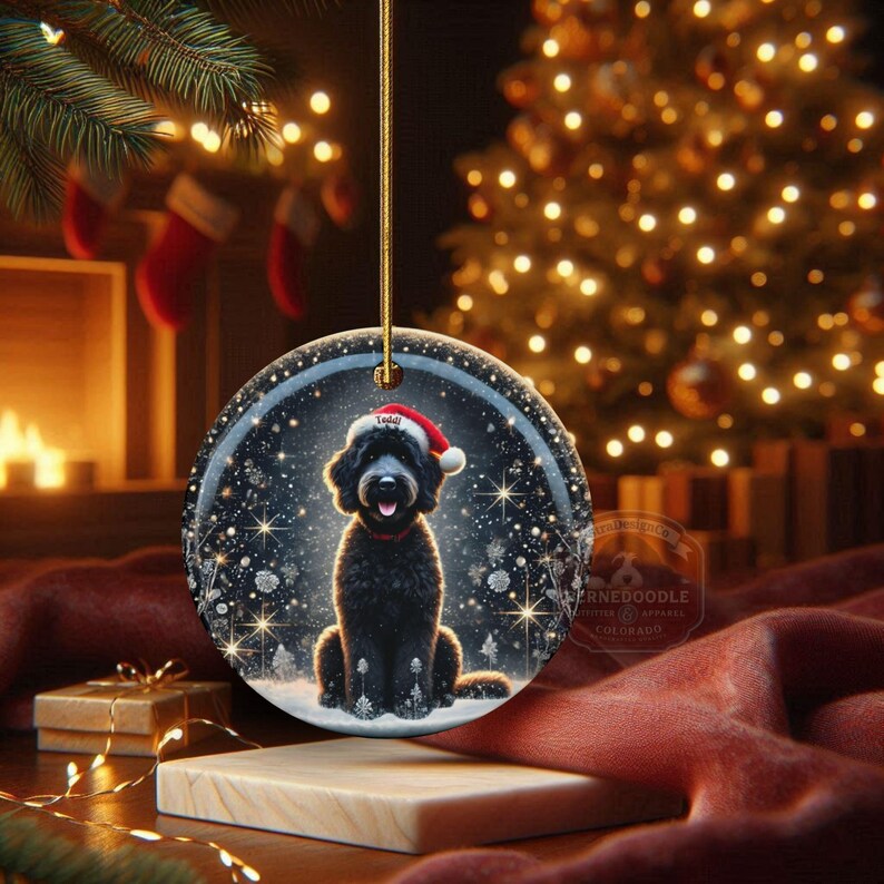May include: Round Christmas ornament featuring a black dog wearing a Santa hat, set against a snowy winter scene with twinkling lights. The ornament is hanging from a gold string, perfect for holiday decor.