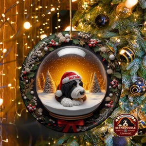 May include: A round Christmas ornament featuring a snow globe design with a Bernedoodle dog wearing a Santa hat that says "Bernie". The ornament is surrounded by a wreath of holly and berries, with a red bow at the bottom. The background shows a Christmas tree.