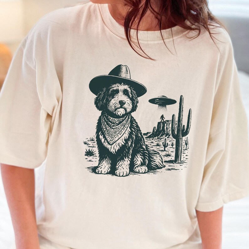 May include: Cream-colored t-shirt featuring a black and white graphic of a dog wearing a cowboy hat and bandana, set against a desert landscape with a UFO. The dog is the central focus, with cacti and a mesa in the background.