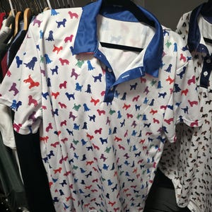 May include: A white short-sleeved polo shirt featuring a repeating pattern of small dog silhouettes in various colours including red, blue, green, and purple. The shirt has a solid blue collar and a button-down placket with two white buttons.