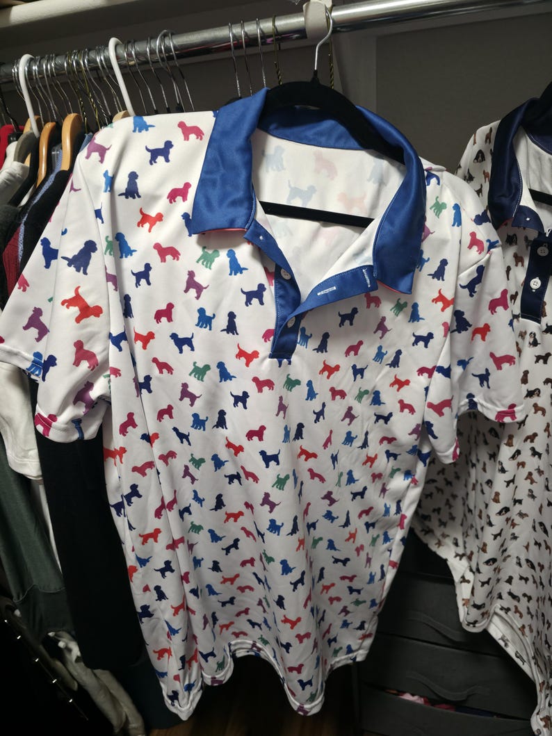 May include: A white short-sleeved polo shirt featuring a repeating pattern of small dog silhouettes in various colours including red, blue, green, and purple. The shirt has a solid blue collar and a button-down placket with two white buttons.
