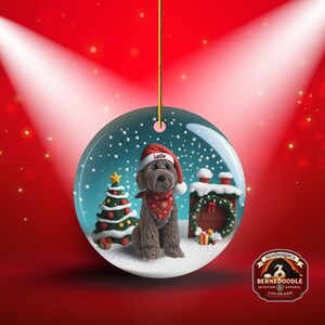 May include: A round Christmas ornament featuring a Bernedoodle dog wearing a Santa hat and bandana. The ornament has a snowy scene with a Christmas tree and a chimney. The dog's hat says "Latte".