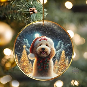 May include: A round Christmas ornament with a gold rim features a smiling dog wearing a Santa hat with the name "Goldie" and a festive scarf. The background shows a snowy forest scene with Christmas trees.