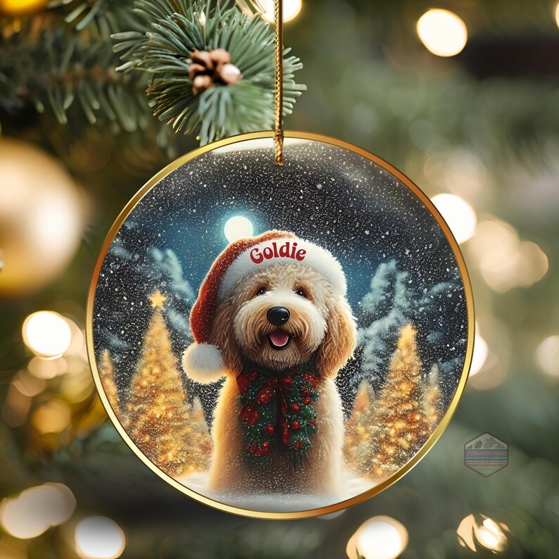 May include: A round Christmas ornament with a gold rim features a smiling dog wearing a Santa hat with the name "Goldie" and a festive scarf. The background shows a snowy forest scene with Christmas trees.