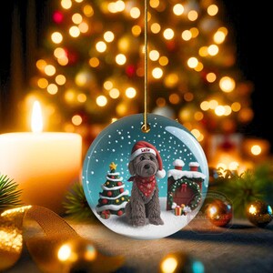 May include: A round Christmas ornament featuring a brown dog wearing a Santa hat and a red bandana. The ornament has a snowy scene with a Christmas tree and a fireplace. The dog's name, "Lalle", is on the hat.
