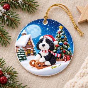 A ceramic ornament featuring a black and white dog wearing a Santa hat and scarf, sitting on a sled and surrounded by a snowy village scene with a Christmas tree and a full moon.