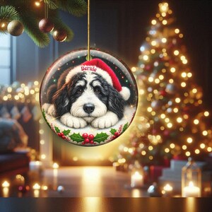 May include: A round Christmas ornament featuring a black and white dog wearing a red Santa hat with the name "Bernie". The ornament is surrounded by a holly and berry design and is hanging from a gold string. The background includes a Christmas tree and lights.