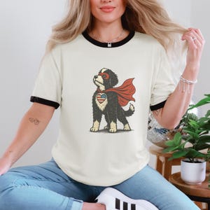May include: Cream-coloured t-shirt with black trim featuring a cartoon dog superhero. The dog has a red cape, a mask, and a heart-shaped emblem with an American flag design. The shirt is a casual style.