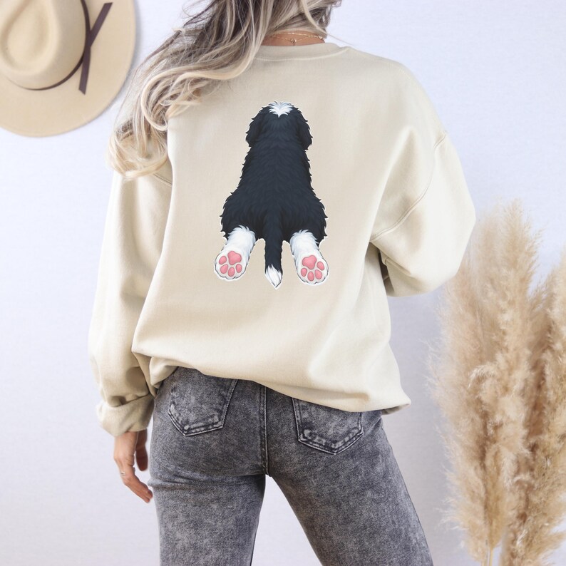Puede incluir: Una sudadera beige con un gr&aacute;fico de la parte trasera de un perro blanco y negro con almohadillas rosas. La sudadera tiene mangas largas y cuello redondo. El gr&aacute;fico del perro est&aacute; centrado en la parte posterior de la sudadera.