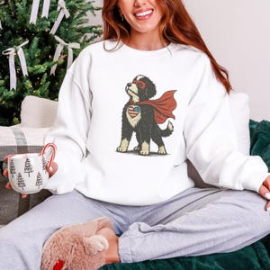 May include: White sweatshirt featuring a cartoon dog dressed as a superhero. The dog has a red cape, a mask, and a heart-shaped emblem with the American flag. The dog is black and white with tan paws.