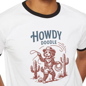 May include: White ringer t-shirt with a black collar and sleeves. The shirt features a cartoon doodle dog dressed as a cowboy, holding a lasso, with the words "HOWDY DOODLE" above the illustration.
