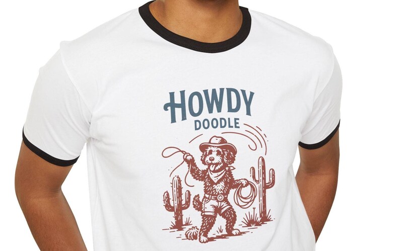 May include: White ringer t-shirt with a black collar and sleeves. The shirt features a cartoon doodle dog dressed as a cowboy, holding a lasso, with the words "HOWDY DOODLE" above the illustration.