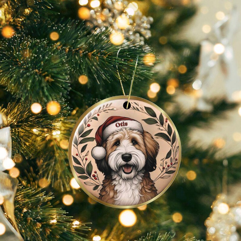 Puede incluir: Un adorno navide&ntilde;o redondo de cer&aacute;mica con un perro marr&oacute;n y blanco que lleva un gorro de Pap&aacute; Noel rojo con el nombre Otis impreso en el borde. El perro est&aacute; enmarcado por una corona circular de hojas verdes y bayas rojas.