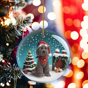 May include: A round Christmas ornament featuring a brown dog wearing a Santa hat with the name "Latte" on it, a red bandana, and a snowy scene. A Christmas tree and a chimney are also visible. The ornament hangs from a gold string.